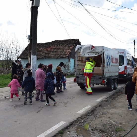 Ein Krankenwagen ist am Straßenrand geparkt und entlädt Kisten. Mehrere Menschen, einschließlich Kinder, warten in der Nähe. Ein Gebäude mit rotem Dach ist im Hintergrund.