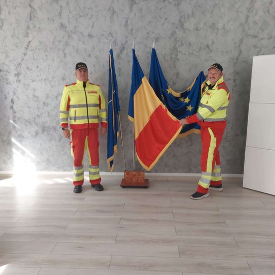 Zwei Männer in reflektierenden Kleidungsstücken stehen vor einer Wand und halten die Flaggen Rumäniens und der Europäischen Union. Sie scheinen an einer formellen Veranstaltung teilzunehmen.