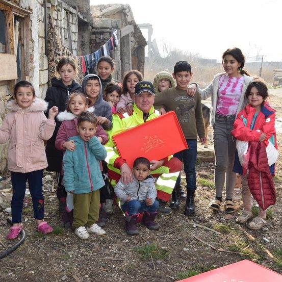Eine Gruppe von Kindern und Erwachsenen, einschließlich eines Mannes in einer reflektierenden Weste, stehen zusammen draußen in der Nähe eines Gebäudes. Sie alle lächeln für ein Foto.