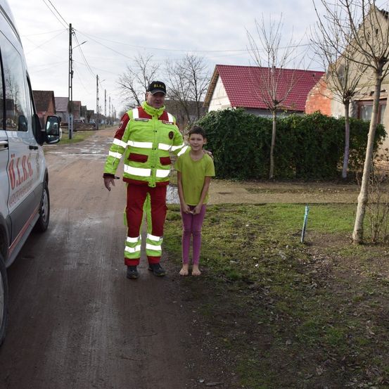 Ein Mann in einer reflektierenden Sicherheitsjacke und ein junges Mädchen in einem grünen Shirt stehen neben einem weißen Van auf einer unbefestigten Straße.
