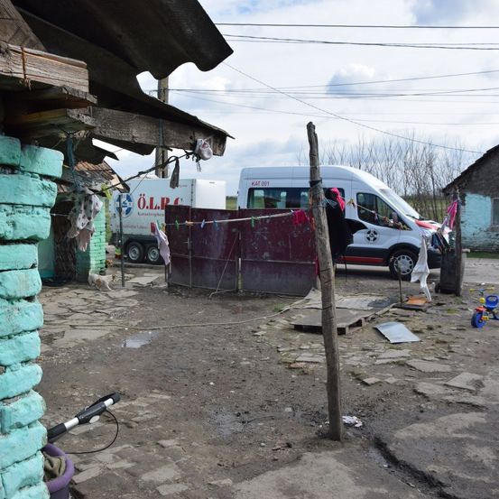 Ein Außenbereich mit einer Holzkonstruktion, einer grünen Ziegelmauer und einer Wäscheleine mit Kleidung. In der Nähe ist ein weißer Lieferwagen mit Text geparkt. Kleidung hängt an der Wäscheleine, und ein kleines Dreirad steht auf dem Boden.