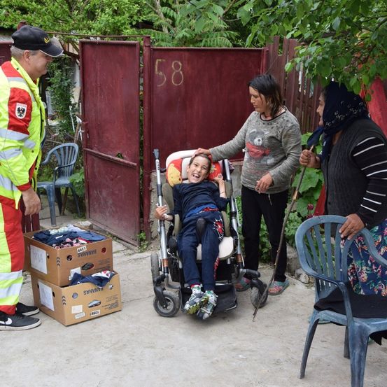 Ein Mann in einer reflektierenden Weste spricht mit einer Frau, die ein Kind im Rollstuhl hält. Zwei Frauen, eine mit einem Stock, stehen in der Nähe. Kartons mit Kleidung liegen auf dem Boden.