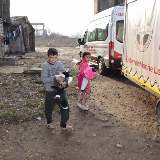 Zwei Kinder gehen neben einem Krankenwagen, ein Junge hält eine Puppe und ein Mädchen einen Rucksack. Dahinter ist ein Krankenwagen geparkt.