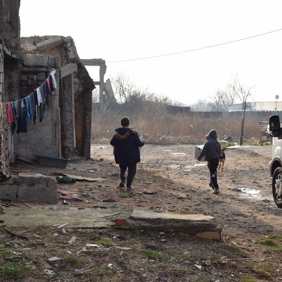 Zwei Personen, eine mit einer Kiste, gehen auf einem unbefestigten Weg an einem beschädigten Gebäude mit einer Wäscheleine vorbei. In der Nähe folgt ein Hund.