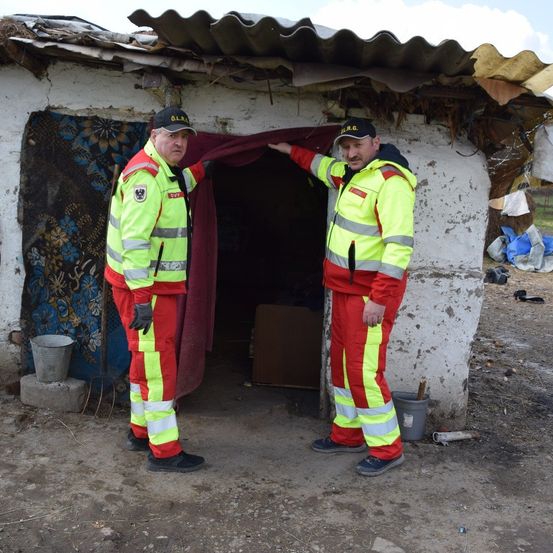 Zwei Männer in reflektierenden Sicherheitsanzügen stehen vor einem beschädigten Gebäude, wobei ein roter Vorhang einen Eingang verdeckt.