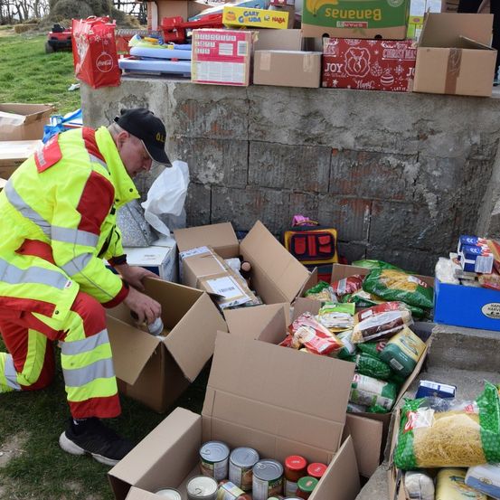Ein Mann in einer Warnschutzkleidung organisiert im Freien Kisten mit Lebensmitteln. Verschiedene Konserven, Nudeln und andere Lebensmittel sind in den Kisten.