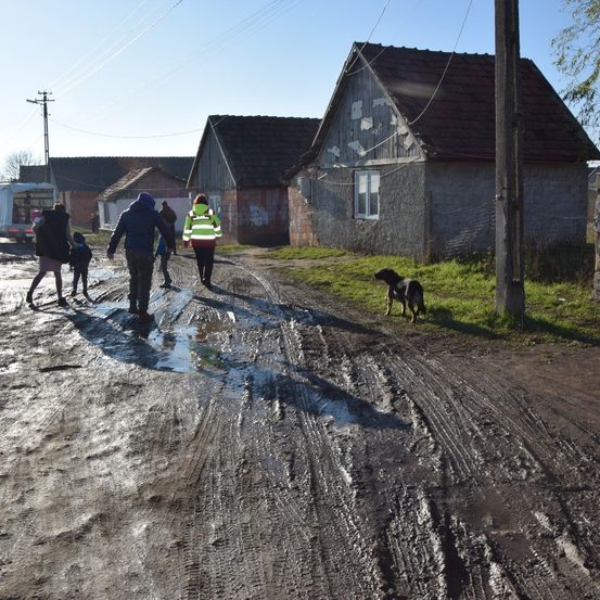 Eine Familie und ein Hund gehen auf einem schlammigen Weg in einem ländlichen Gebiet, mit Häusern und einem Lieferwagen im Hintergrund.