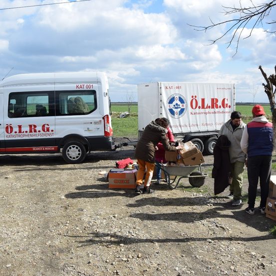 Zwei weiße Fahrzeuge mit den Buchstaben O.L.R.G. sind auf einem Erdweg geparkt. Menschen entladen Kisten aus einem der Fahrzeuge. Eine Person schiebt einen mit Kisten gefüllten Schubkarren.