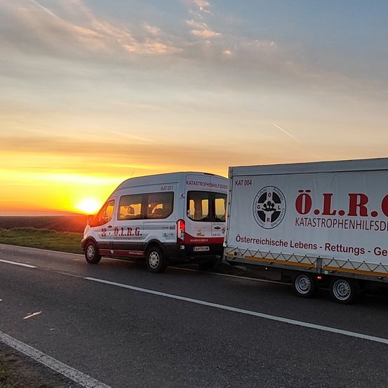 Ein weißer Van und ein Anhänger mit Notfalldienstmarkierungen fahren bei Sonnenuntergang auf einer Straße.