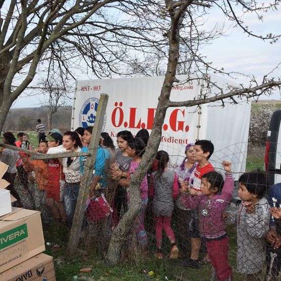 Eine Gruppe von Kindern und Erwachsenen wartet hinter einem Zaun in der Nähe eines weißen Lastwagens mit der Aufschrift O.L.J.G. Sie befinden sich auf einem Rasen mit Pappkartons in der Nähe.