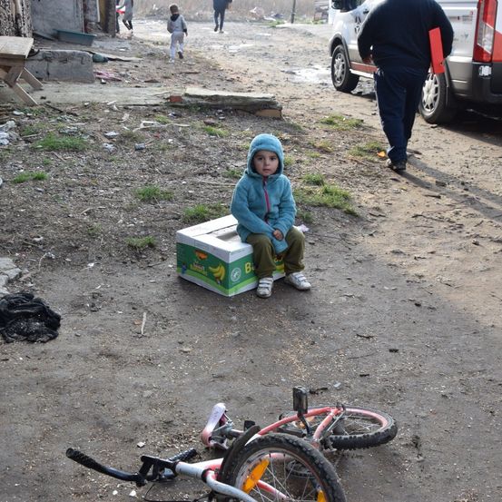 Ein Kind sitzt auf einem Karton auf einem unbefestigten Weg, ein Fahrrad daneben. Im Hintergrund geht eine Person, und ein Fahrzeug ist geparkt.