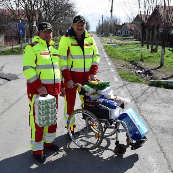 Zwei Männer in gelben Sicherheitswesten stehen neben einem mit Paketen beladenen Rollstuhl. Sie befinden sich auf einer Straße mit Häusern und Bäumen im Hintergrund.
