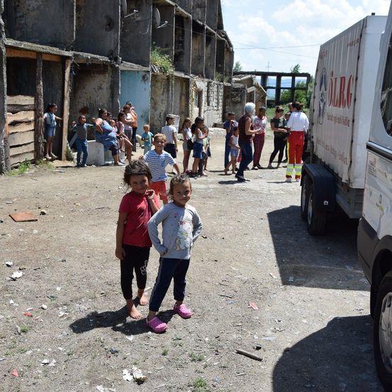 Zwei Kinder stehen auf einer Straße vor einem alten Gebäude mit Menschen um sie herum, in der Nähe eines Lastwagens.