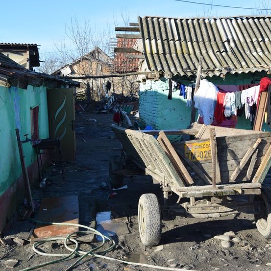Eine schmale Straße mit einem Holzwagen und einem Gebäude, an dem Kleidung hängt. Der Wagen hat ein Nummernschild, das 'TM-026' liest.