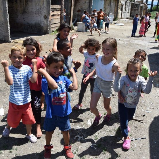 Mehrere Kinder stehen in einer Reihe, lächeln und posieren für ein Foto in einem Außenbereich mit schmutziger Erde und Gebäuden im Hintergrund.