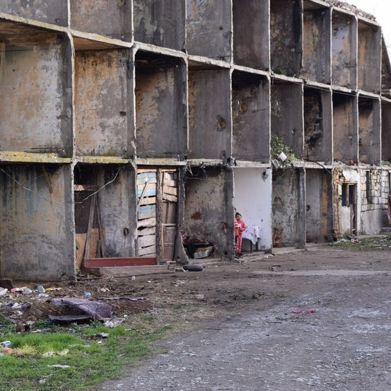 Ein verfallenes mehrstöckiges Gebäude mit stark beschädigten und mit Graffiti bedeckten Außenwänden steht an einer vernachlässigten Straße. Ein Kind steht bei einem Tisch mit einem weißen Tuch, während eine Person in der Nähe sitzt.