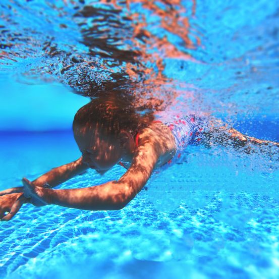 Ein junges Mädchen schwimmt unter Wasser in einem Pool, die Arme vor sich ausgestreckt. Das Wasser ist klar und blau.