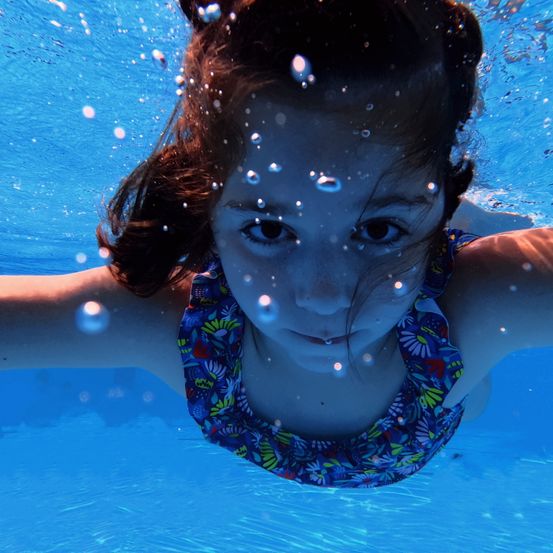 Ein junges Mädchen schwimmt unter Wasser in einem Pool, umgeben von Blasen, in einem Blumenbadeanzug.