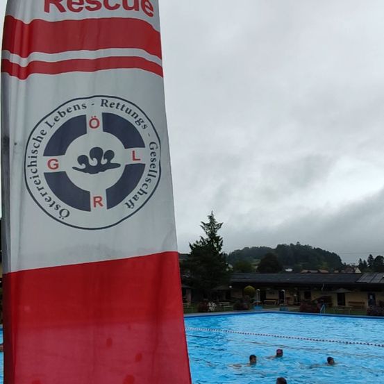 Eine Rettungsschwimmerflagge steht in roten Buchstaben 'Rettung'. Ein Schwimmbecken mit Schwimmern und einem bewölkten Himmel im Hintergrund.
