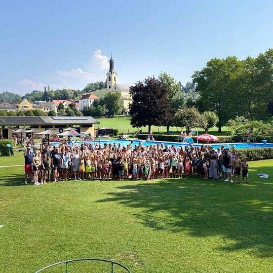 Eine große Gruppe von Menschen versammelt sich an einem sonnigen Tag vor einem Schwimmbad, mit einer Kirche und Hügeln im Hintergrund.