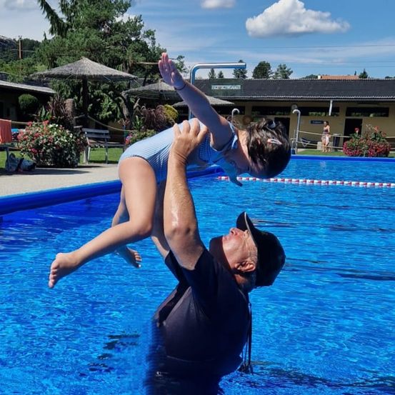 Ein Mann hält ein Mädchen in einem Badeanzug in einem Schwimmbecken. Dahinter befindet sich ein Gebäude mit Terrasse, Sonnenschirmen und Blumen.