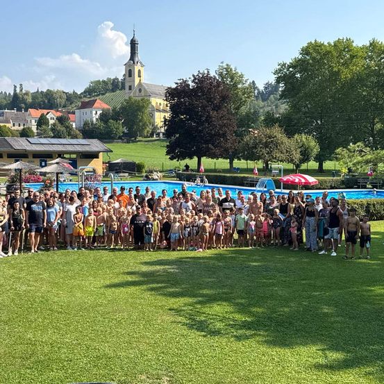Eine große Gruppe von Menschen versammelt sich draußen in der Nähe eines Pools, mit einer Kirche und Häusern im Hintergrund. Bäume und ein roter Regenschirm sind sichtbar.
