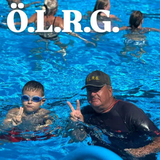 Mehrere Menschen schwimmen im Pool. Eine Person macht ein Friedenszeichen mit den Händen. Die Buchstaben O.L.R.G. sind oben angezeigt.