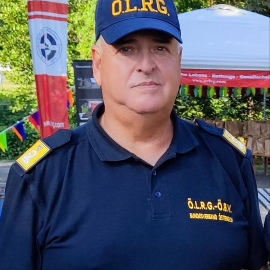 Ein Mann in einer dunkelblauen Uniform mit gelber Schrift und einer blauen Mütze mit den Buchstaben O.L.R.G. steht vor einem weißen Zelt.