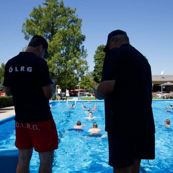 Zwei Männer in schwarzen T-Shirts und roten Shorts mit 'OLRG' stehen am Pool. Mehrere Menschen schwimmen im Pool. Im Hintergrund befinden sich Bäume und ein Gebäude.