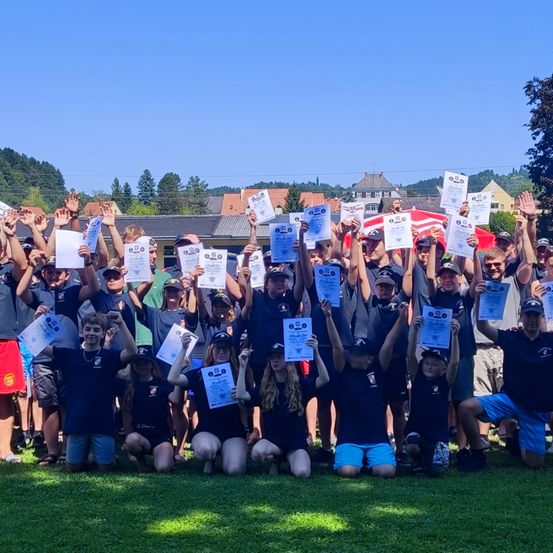 Eine Gruppe von Menschen, wahrscheinlich ein Team, posiert für ein Foto auf einem Rasenfeld, hält Urkunden und trägt Uniformen. Im Hintergrund befinden sich Gebäude und Bäume.