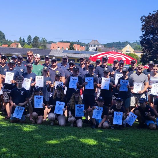 Eine Gruppe von Menschen in einheitlichen Kleidungsstücken posiert an einem sonnigen Tag für ein Foto, hält Urkunden in der Hand und lächelt. Hinter ihnen steht ein rotes Zelt und eine malerische Aussicht auf Häuser und Bäume.