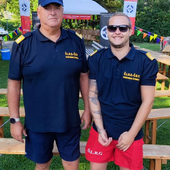 Zwei Männer in gleichen Uniformen stehen auf einer Bank und lächeln für ein Foto. Der Mann auf der linken Seite trägt eine Uhr und eine Kappe. Hinter ihnen sind Bänke, ein Zelt und ein Baum.
