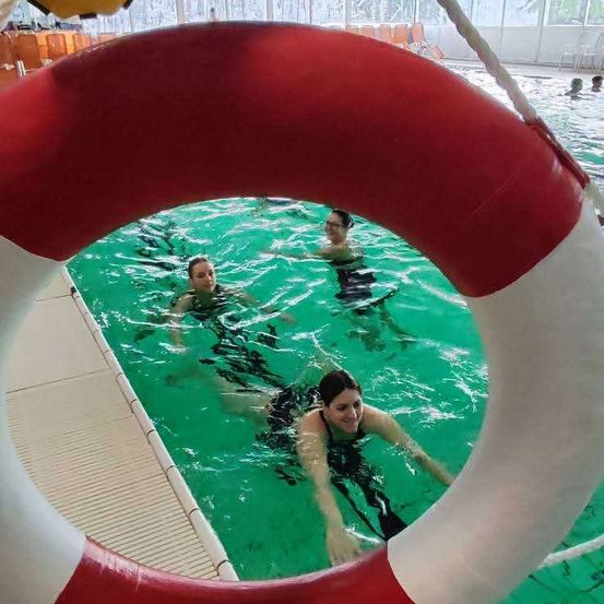 Drei Frauen schwimmen in einem Pool mit einem roten Rettungsring im Vordergrund. Der Pool ist grün, und es gibt Menschen im Hintergrund.