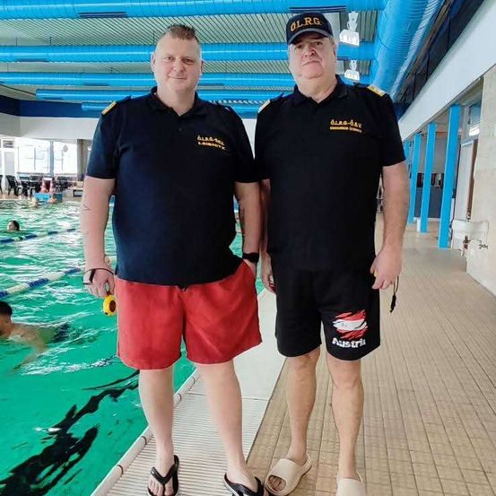 Zwei Männer in schwarzen Uniformen und Mützen stehen neben einem Pool, einer hält eine Pfeife. Sie schauen in die Kamera.