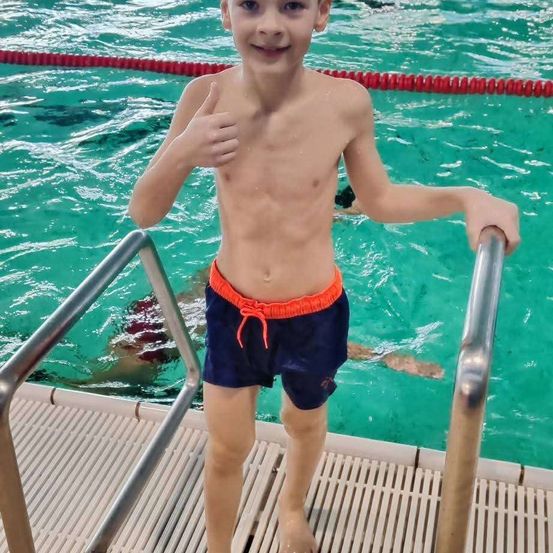 Ein Junge steht mit einem Lächeln auf der Poolbank und gibt einen Daumen hoch, während ein anderer im Hintergrund schwimmt.