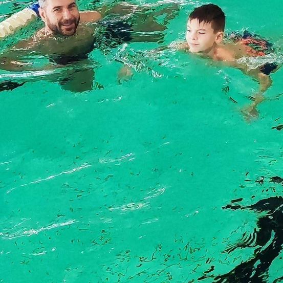 Ein Mann und ein Junge schwimmen im Pool, lächeln und schauen zur Kamera. Der Junge hat die Augen geschlossen.