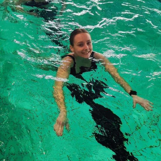 Eine Frau schwimmt in klarem, grünem Wasser. Sie trägt einen schwarzen Badeanzug und sieht glücklich aus. Sie hat ein Armband am linken Handgelenk.