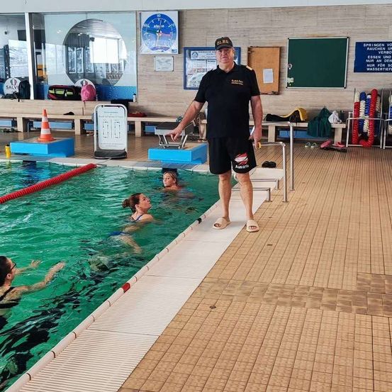 Ein Mann in einer schwarzen Uniform steht am Rand eines Schwimmbeckens und beobachtet einige Schwimmer beim Üben. Das Becken hat eine blaue Bahnenlinie.