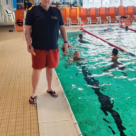 Ein Mann steht neben einem Hallenbad und hält eine Pfeife. Mehrere Menschen schwimmen im Pool.