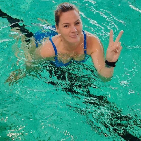 Eine Frau in einem blauen Badeanzug schwimmt in einem Pool und macht ein Friedenszeichen mit den Händen. Sie sieht glücklich und entspannt aus. Das Wasser ist klar, und der Pool ist gut beleuchtet.