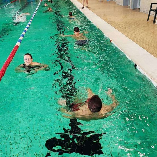 Mehrere Menschen schwimmen in einem Schwimmbecken mit Bahnlinien. Eine Frau trägt eine Brille und ein schwarzes Badeanzug. Ein Mann schwimmt mit dem Gesicht nach unten. Das Becken hat einen gefliesten Boden.