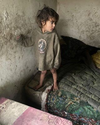 Ein kleines Kind steht barfuß in einem Zimmer und trägt eine grüne Kapuzenjacke. Die Wände sind verschmutzt, und im Hintergrund befindet sich ein Bett mit einem farbenfrohen Quilt.
