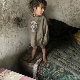 Ein kleines Kind steht barfuß in einem Zimmer und trägt eine grüne Kapuzenjacke. Die Wände sind verschmutzt, und im Hintergrund befindet sich ein Bett mit einem farbenfrohen Quilt.