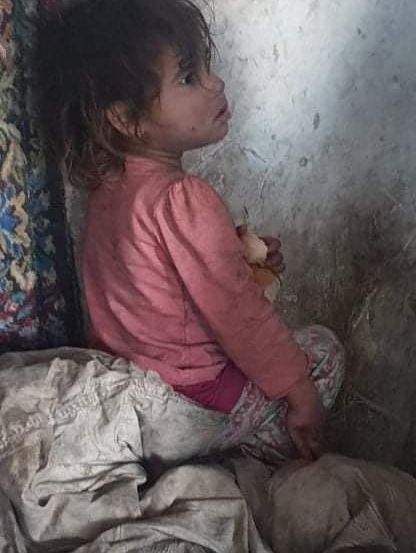 Ein kleines Mädchen sitzt auf dem Boden mit dem Rücken an einer Wand und hält ein Stück Essen in der Hand. Sie schaut nach oben, möglicherweise wartet oder beobachtet sie etwas. Sie trägt ein rosa Langarmshirt und Hose.