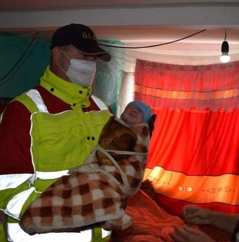 Ein Mann in einer Sicherheitsweste und Gesichtsmaske hält ein Baby, das in eine Decke gewickelt ist. Der Raum hat einen roten Vorhang und eine Glühbirne an der Decke.