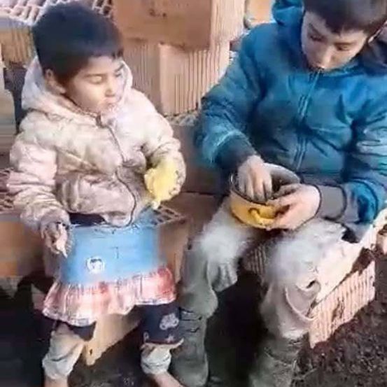 Zwei Kinder sitzen auf einem Stapel Ziegelsteine, eines in einem blauen Mantel und das andere in einem rosa Mantel.