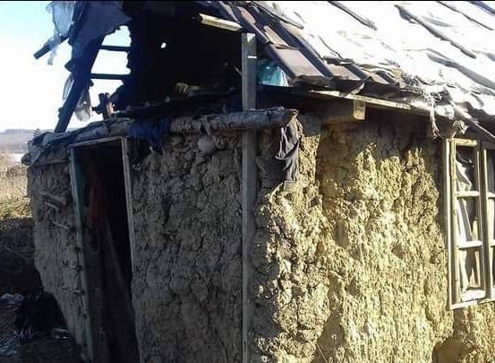 Ein beschädigtes Haus mit einem kaputten Dach und offener Tür steht in einem ländlichen Gebiet. Das Äußere besteht aus Lehm und weist sichtbare Abnutzungserscheinungen auf.