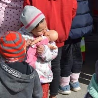 Ein junges Mädchen hält eine Puppe und steht zwischen anderen Menschen. Sie trägt eine Mütze und rote Hosen. Ein weiteres Kind in einem roten Hut schaut sie an. Hinter ihnen stehen Erwachsene in verschiedenen farbigen Kleidungsstücken.
