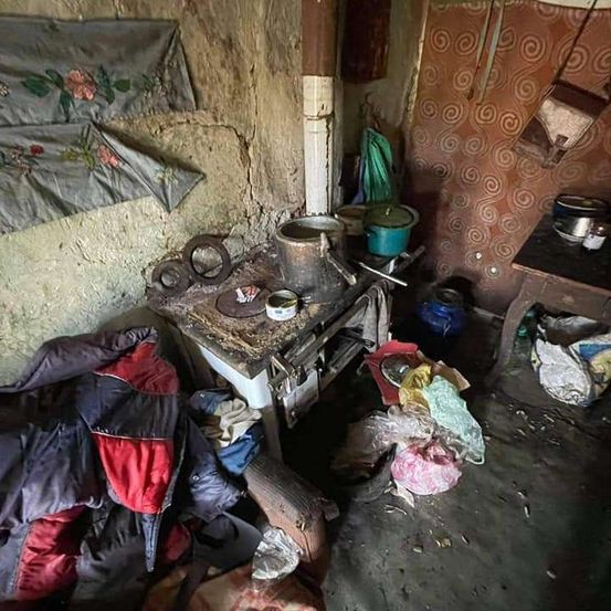 Ein unordentliches Zimmer mit einem kleinen Ofen, Töpfen und auf dem Boden verstreuten Tüten. Es gibt eine Wand mit Tapeten und hängenden Gegenständen.