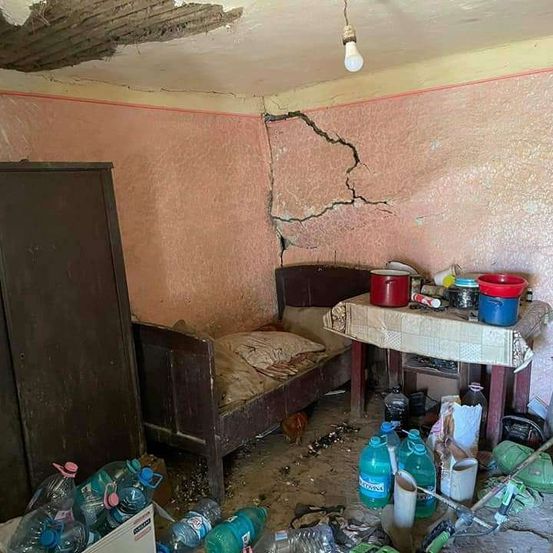 Ein kleiner Raum mit beschädigten Wänden, einer rissigen Decke und einem Bett. Ein Tisch hat verschiedene Gegenstände, darunter einen Eimer, eine Flasche und einen Topf. Mehrere Plastikflaschen sind auf dem Boden verstreut.